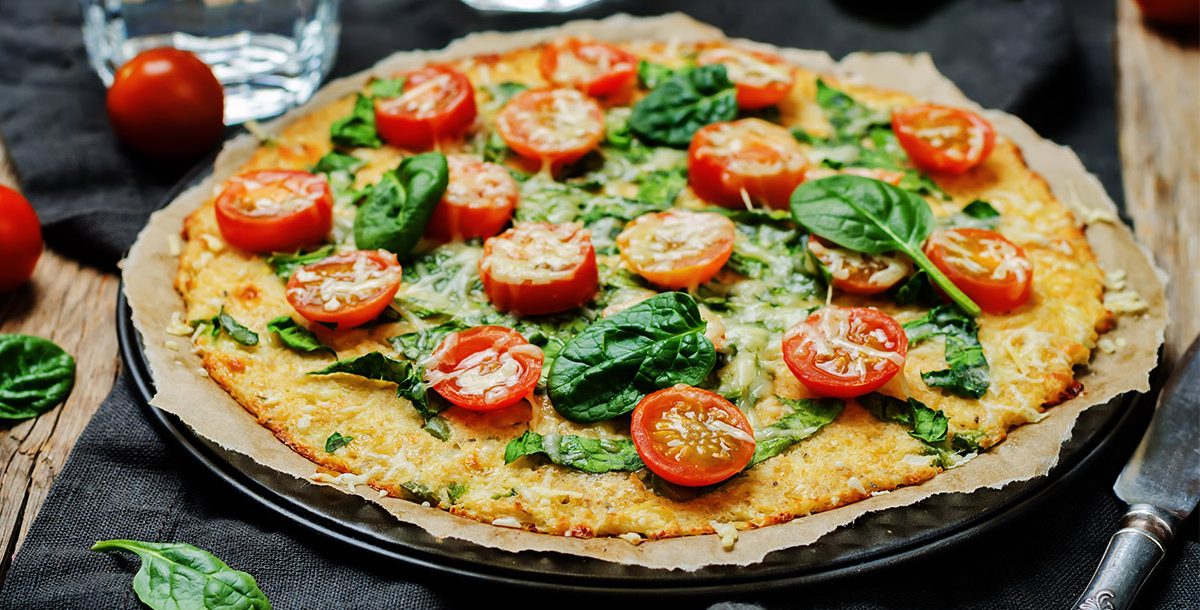 Cauliflower Crust Pizza sitting on a table with spinach and tomatoes
