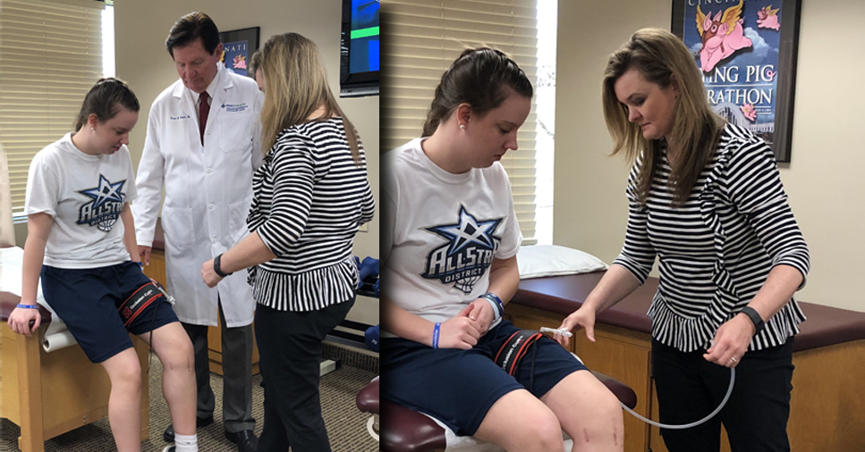 A doctor in a white coat and a woman in a striped shirt assist a young female patient with fitting a blood pressure cuff around her knee