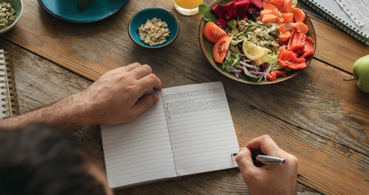A man writing in a notebook about healthy eating rules for weight loss on a wooden table with a colorful salad in a bowl next to a small bowl of nuts