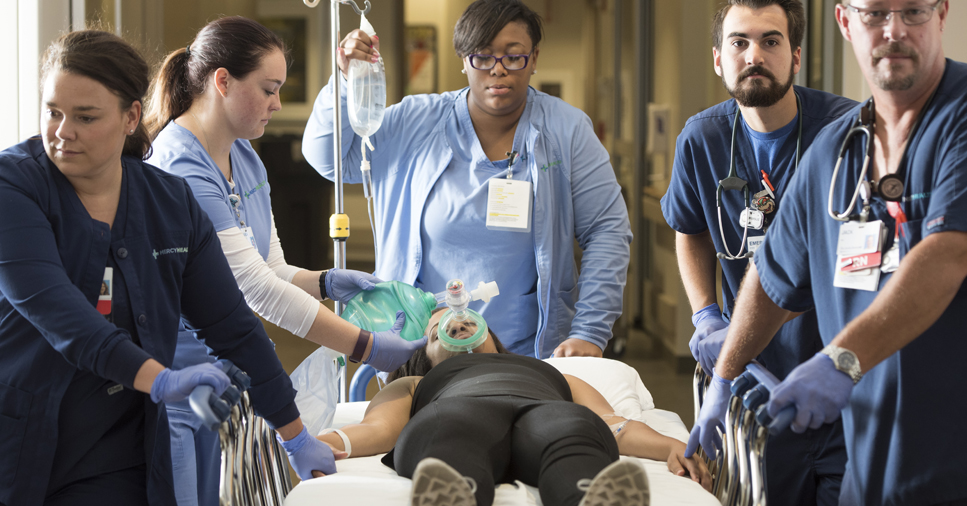 A team of five nurses in blue scrubs attend to a female patient who is unresponsive and laying in a hospital bed