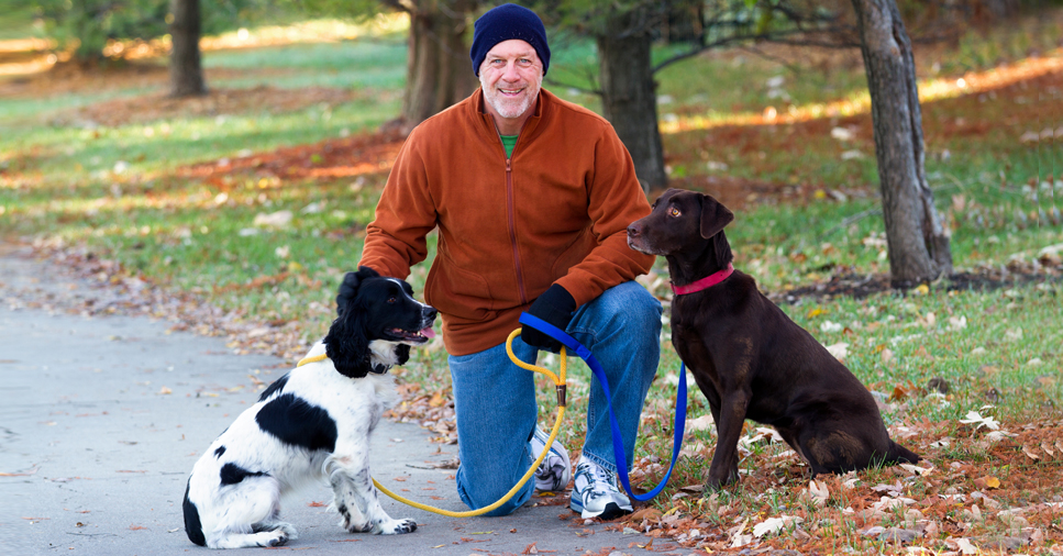 Middle-aged man wearing a brown jacket and jeans kneeling down to pet his two dogs on a fall day. One dog is brown and the other is white with black spots.