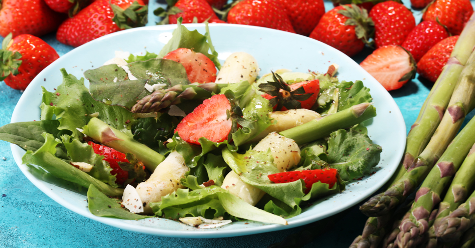 A salad with spring vegetables and fruits such as strawberries and asparagus on a white plate sits on a table